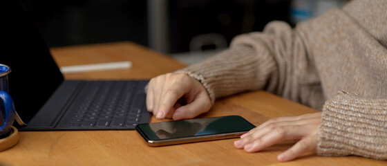 Female touching on smartphone while sitting at workspace with digital tablet on wooden table