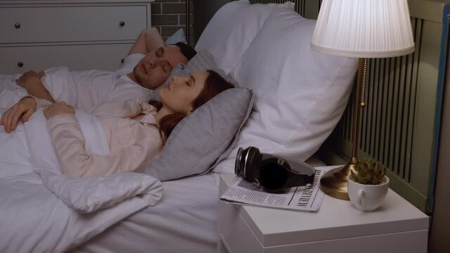 Young irritated woman putting headphones on because of snoring husband in bed