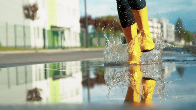 SLOW MOTION, LOW ANGLE, CLOSE UP, DOF: Cinematic shot of a girl running across a crystal clear puddle after an intense rainstorm. Happy woman wearing yellow rain boots runs across a glassy puddle.
