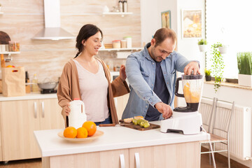 Cheerful couple prepares smoothie using blender. Wife holding milk bottle in kitchen. Healthy carefree and cheerful lifestyle, eating diet and preparing breakfast in cozy sunny morning