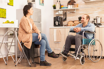 Fototapeta na wymiar Disabled man in wheelchair and wife smiling at each other in kitchen. Disabled paralyzed handicapped man with walking disability integrating after an accident.