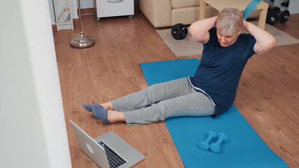 Sportive senior woman on yoga mat exercising looking at online fitness program. Old person pensioner online internet exercise training at home sport activity at elderly retirement age
