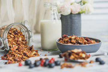 Homemade granola or muesli made of oats, nuts, honey, cinnamon, fruits in blue plate on white table with milk and flowers. Beautiful healthy snack on breakfast. Copy space.