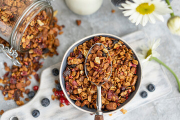 Homemade granola or muesli made of oats, nuts, honey, cinnamon, fruits on white table with milk and flowers. Beautiful healthy snack on breakfast. Top view, flat lay, copy space.