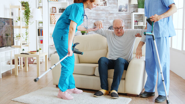 Team Of Nurses Or Social Workers Helping An Old Disabled Man To Walk With His Crutches Out Of The Nursing Home Room.