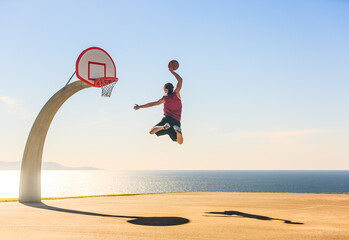 Basketball player jumping in the air scoring an amazing energetic slam dunk at the street ball court with a beautiful ocean view in the background. Sports, motivation and elevation.