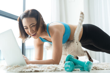 Asian woman in sportwear practicing yoga and watching tutorial lesson on laptop doing exercise in living room with a cat at home