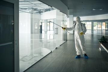 A man wearing disinfection suit spraying with sanitizer the glass doors' handles in an empty shopping mall to prevent covid-19 spread. Health awareness, clean, defence concept.