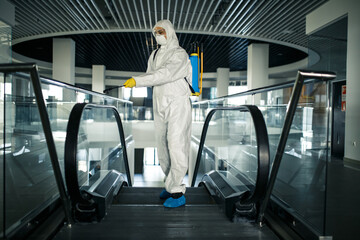 Fototapeta na wymiar Disinfection worker wearing a protective suit professionally cleans up an escalator in an empty business center. A man equiped with antibacterial clothes sterilizes shopping mall. Covid concept.