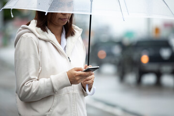 Fototapeta premium Asian woman is using on smartphone, checking social media network and holding umbrella while waiting taxi on the city street in the rainy day.