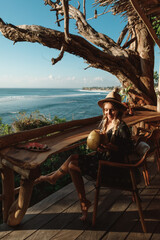 Fototapeta na wymiar Happy blonde woman in hat on the tropical beach sitting in cafe with coconut