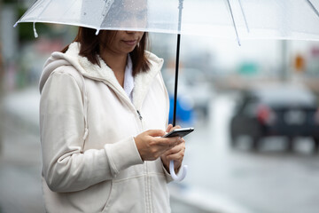 Asian woman is using on smartphone, checking social media network and holding umbrella while waiting taxi on the city street in the rainy day.