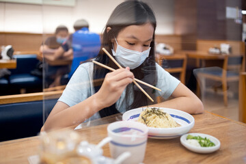 Asian child girl wear face mask with food,plastic shield as a barrier partition on the tables in...