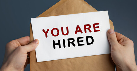 Male hands holding craft envelope with text YOU ARE HIRED on blue background