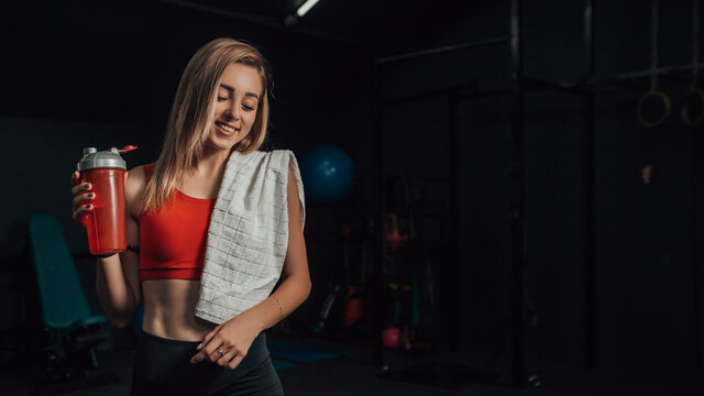Portrait of young smiling girl after productive training standing in gym or fitness center. Towel on shoulder, bottle of water in hand. Health care concept. 