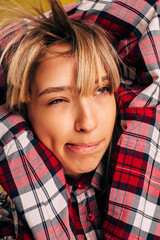 Weird funny young blonde woman blowing up on her bangs during posing on camera. Girl in red checkered shirt hold hands above head. Clothing advertisement.