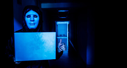 Anonymous computer hacker in white mask and hoodie. Obscured dark face holds a laptop tight in his hands on target room background, Data thief, internet attack, darknet and cyber security concept
