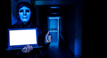 Anonymous computer hacker in white mask and hoodie. Obscured dark face holds a laptop tight in his hands on target room background, Data thief, internet attack, darknet and cyber security concept