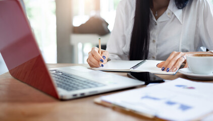 Close-up of hand businesswoman writing on notepad  while she working with computer laptop and smart phone on desk