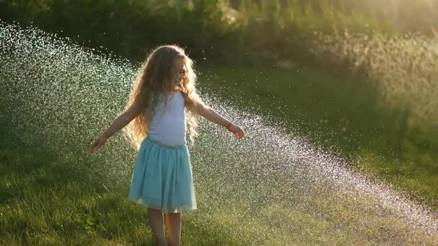 A little girl is having fun on the green lawn, which is poured with a stream of water in summer. Cooling in hot summer under running water on the lawn.