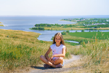 Fototapeta na wymiar A little girl a child with long hair sits on a path with a mountain landscape with rivers and green meadows