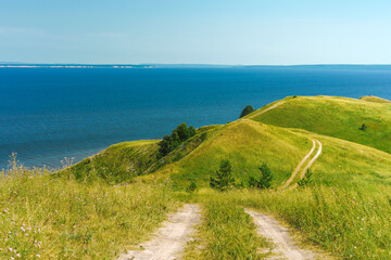 A wide shot of a curved gravel road through a lush green hillside with the sea or lake in the background.