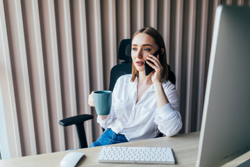 Cute young woman with on phone while working on laptop computer at desk next to coffee cup with...