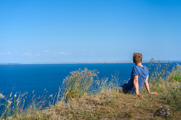 Fototapeta na wymiar A blond man in a blue shirt sits on a cliff overlooking the blue sea and Islands