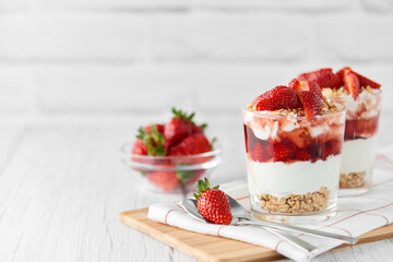 Homemade layered dessert with fresh strawberries, cream cheese or yogurt, granola and strawberry jam in glasses on white wood background. Healthy organic breakfast or snack concept. Selective focus.