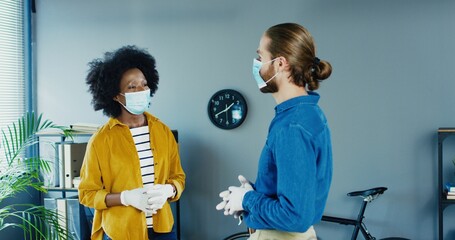 Mixed-race male and female office workers in masks and gloves having conversation in cabinet. Pretty African American woman chatting with Caucasian man at work in quarantine. Working place concept