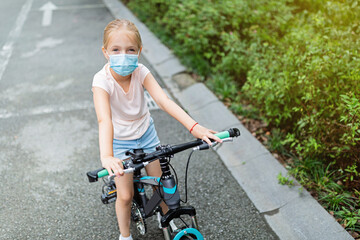 Active school kid girl in medical mask riding a bike on summer sunny day. Happy child biking during coronavirus covid-19 epidemic.