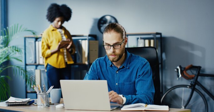 Handsome Caucasian male worker typing on laptop and writing down in notebook while sitting at office. African American girl walking on background and tapping on tablet in cabinet. Mixed-races concept