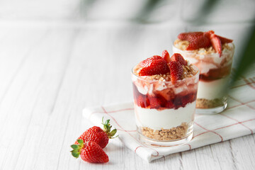 Homemade layered dessert with fresh strawberries, cream cheese or yogurt, granola and strawberry jam in glasses on white wood background. Healthy organic breakfast or snack concept. Selective focus.