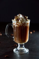 Close up of a glass cup of coffee with whipped cream and shaved chocolate on it and roasted coffee beans on dark background. Concept of ready to eat food, tasty snack. Selective focus, copy space.