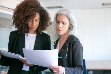 Professional female partners reading document in office room. Two beautiful successful content businesswomen studying documentation report together. Teamwork, business and management concept