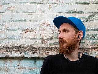 Fototapeta premium Portrait of young red bearded man in black t-shirt and snapback in rock style on urban background listening to music with headphones. Hipster guy in wireless earphones on old blue brick wall backdrop.