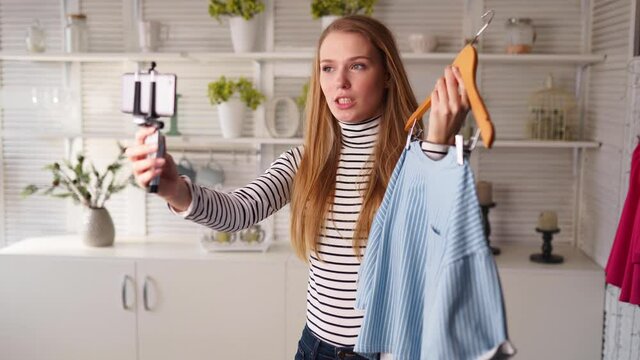 Fashion blogger woman in turtleneck showing casual colorful shirts on smartphone camera. Stylist influencer girl with trendy clothes filming vlog episode for her channel. Opinion leader sets trends.