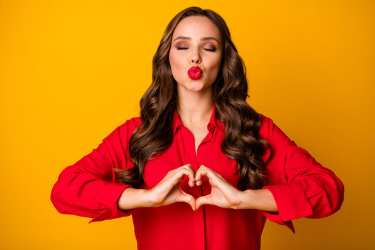 Photo of attractive pretty wavy lady hold hands fingers making heart figure symbol romantic mood send air kisses eyes closed wear red shirt isolated yellow bright color background