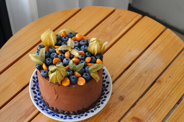 On a round wooden table is a homemade chocolate cake decorated with physalis and blueberries...