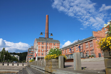 Wooden embankment with steps and flower beds against the background of a red brick factory with a large chimney on the shore of lake Vesiyarvi in the city of Lahti. Finland.
