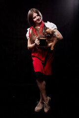 Girl in national ethnic clothing with small dog on hands posing in a Studio on black background. A young woman in a red dress and white shirt with long hair and ponytails and her pet Brussels Griffon.