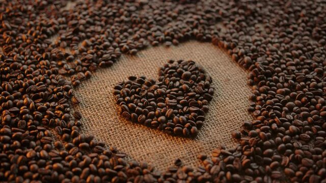 Woman's hand form heart shape illustration made of roasted arabica coffee grains. Coffee beans and autentic burlap background. Dolly shot. Warm coloured