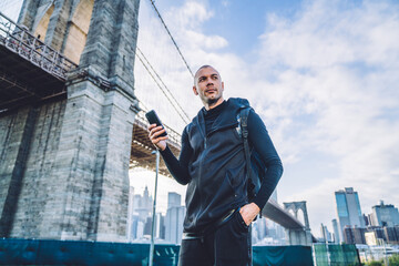 Young caucasian man standing near Brooklyn bridge and using navigation application on mobile phone....