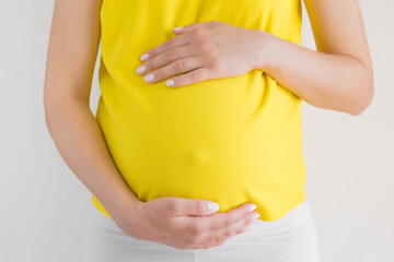 Young woman in yellow clothes touching big belly with hand. Isolated on gray background. Emotional...