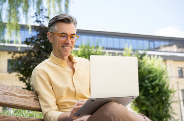 Freelancer copywriter using laptop computer, typing on keyboard. Portrait of handsome mature man working start up project, sitting on bench. Successful business. Smiling man having video conference