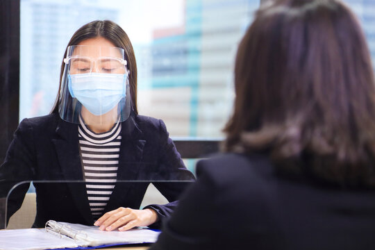 Manager From HR Department Wearing Facial Mask Is Interviewing New Applicant And Reading Her Resume And Profile Through The Partition For Social Distancing And New Normal Policy