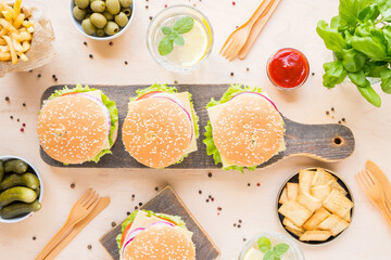 Dinner table with burger, french fries, vegetables, sauces, snacks and lemonade on wooden background, top view