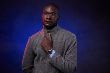 Stylish African American man in gray jacket and glasses on dark background with blue light. Studio shot