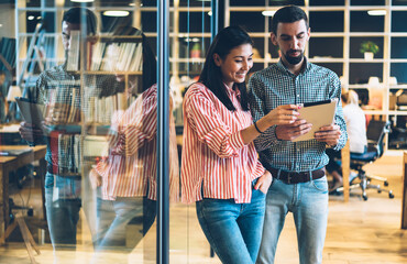 Smiling young woman viewing common photos on modern digital tablet standing with male colleague on break in office,positive employees satisfied with good results of marketing strategy using touchpad.