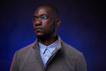 Stylish African American man in gray jacket and glasses on dark background with blue light. Studio shot